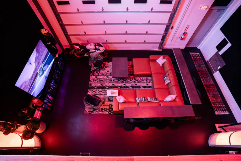 Aerial overhead view of Sanctum Studio lounge in Roanoke TX showing red leather sectional, patterned area rug, large TV display, motorcycle prop, and roll-up garage door