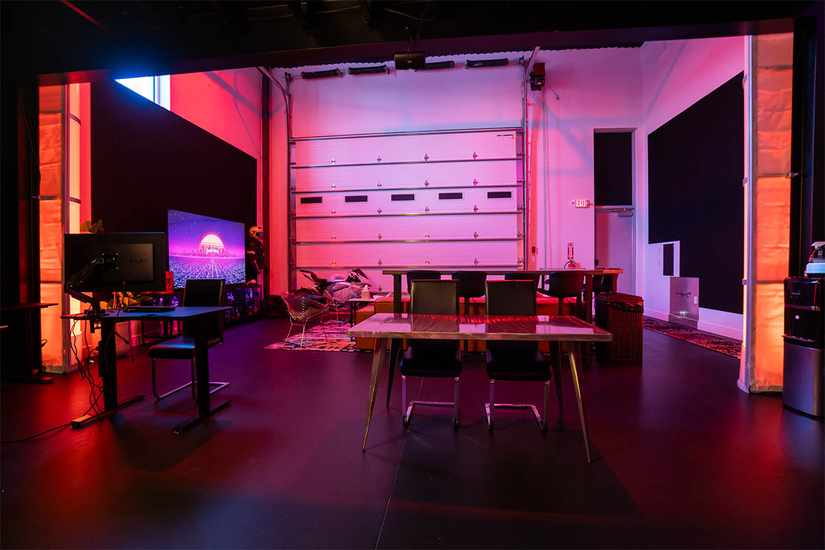 Sanctum Studio ground floor with standing desk workstation, conference table, roll-up garage door, and large display screen lit in pink and red in Roanoke TX
