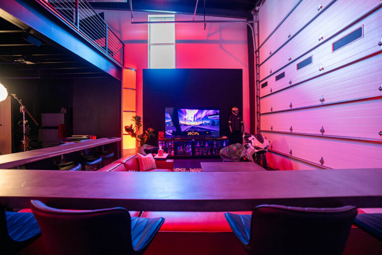 Sanctum Studio lounge area viewed from bar seating with large TV display, red leather sectional, motorcycle prop, and roll-up garage door in Roanoke TX