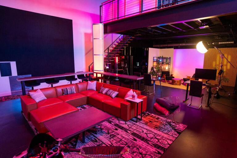 Sanctum Studio lounge and filming zone with red leather sectional, interview chairs, studio light, acoustic slat wall, and pink-red ambient LED lighting in Roanoke TX